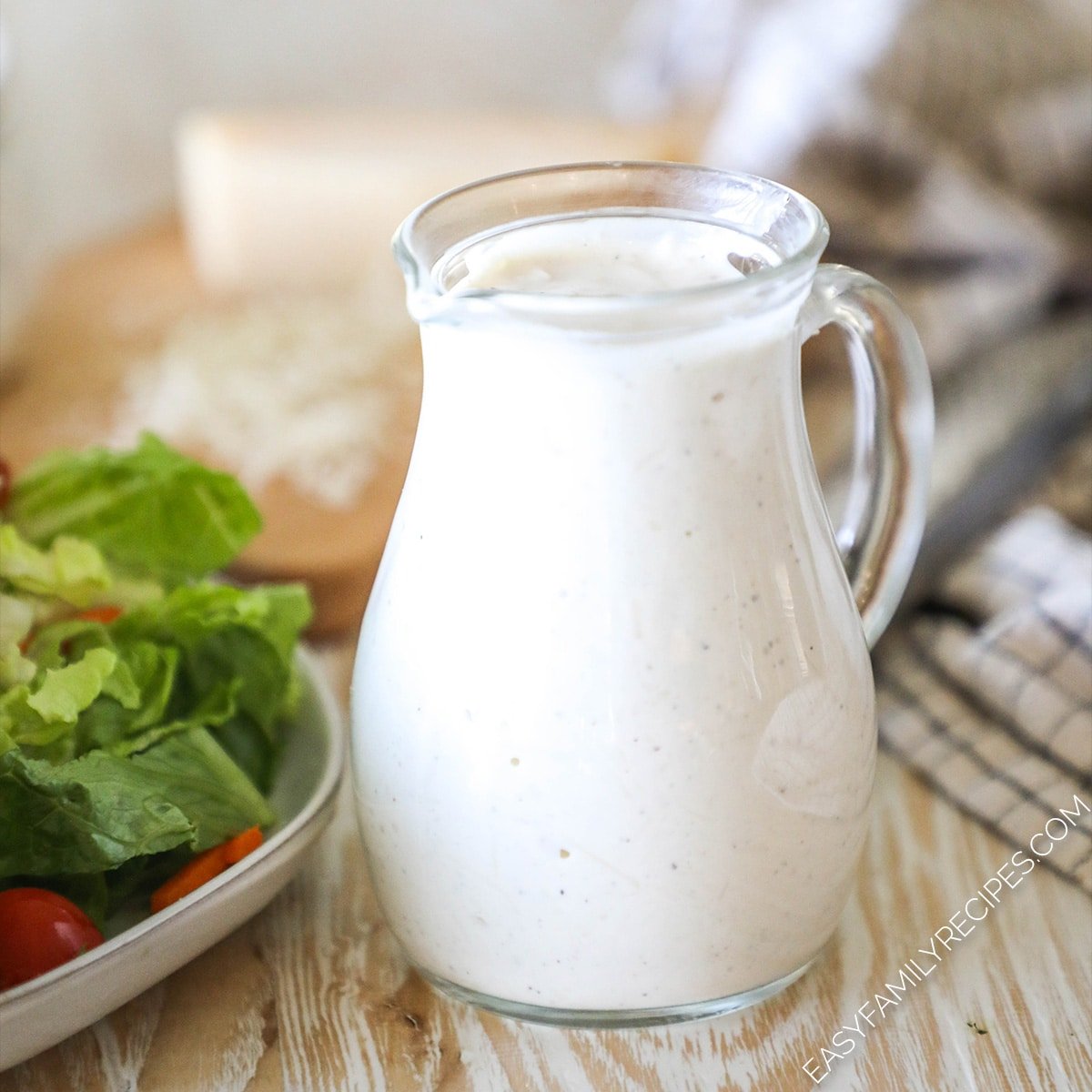 a pitcher of creamy dressing next to a plate with salad.